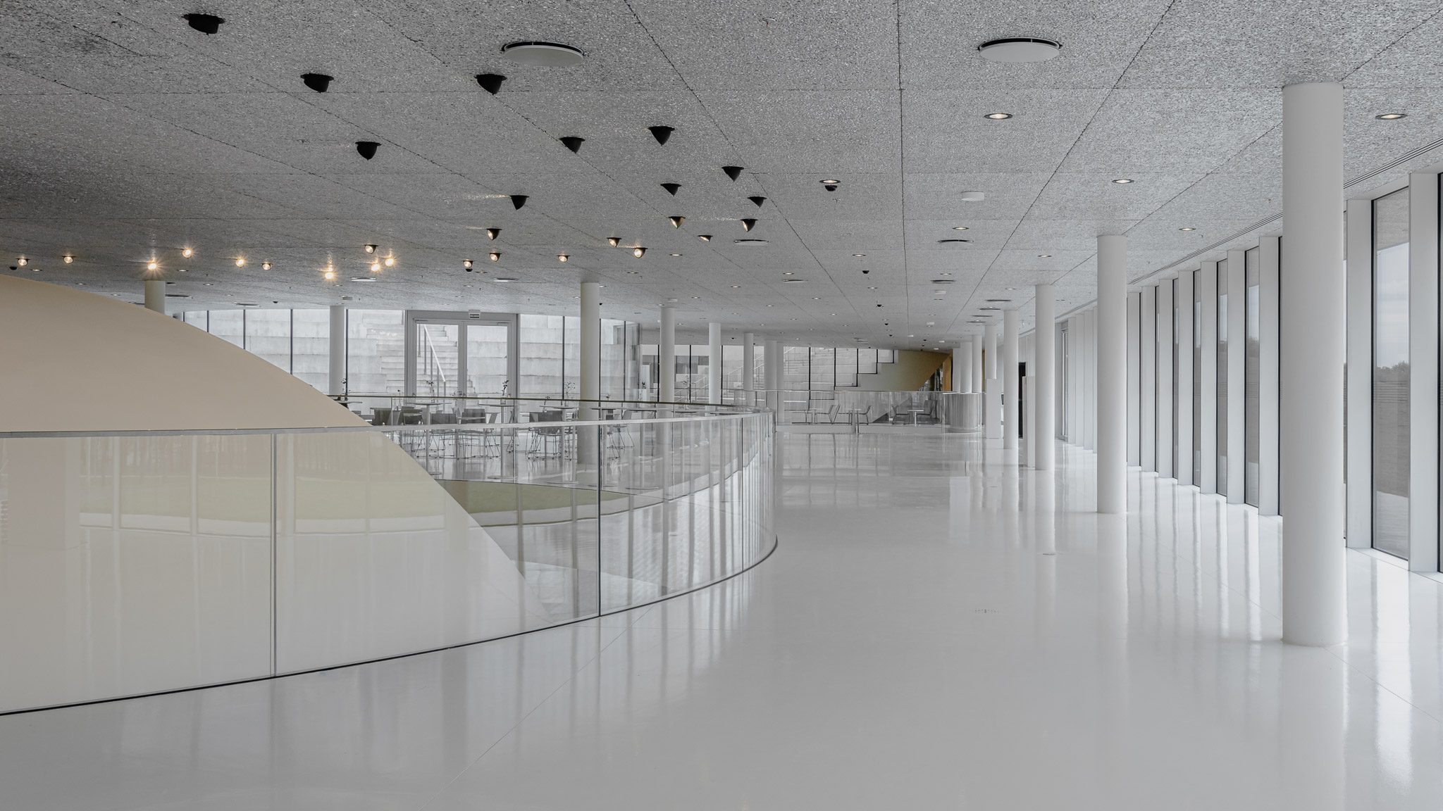 White gallery with a curved glass balustrade and suspended lights, Mokslo Sala
